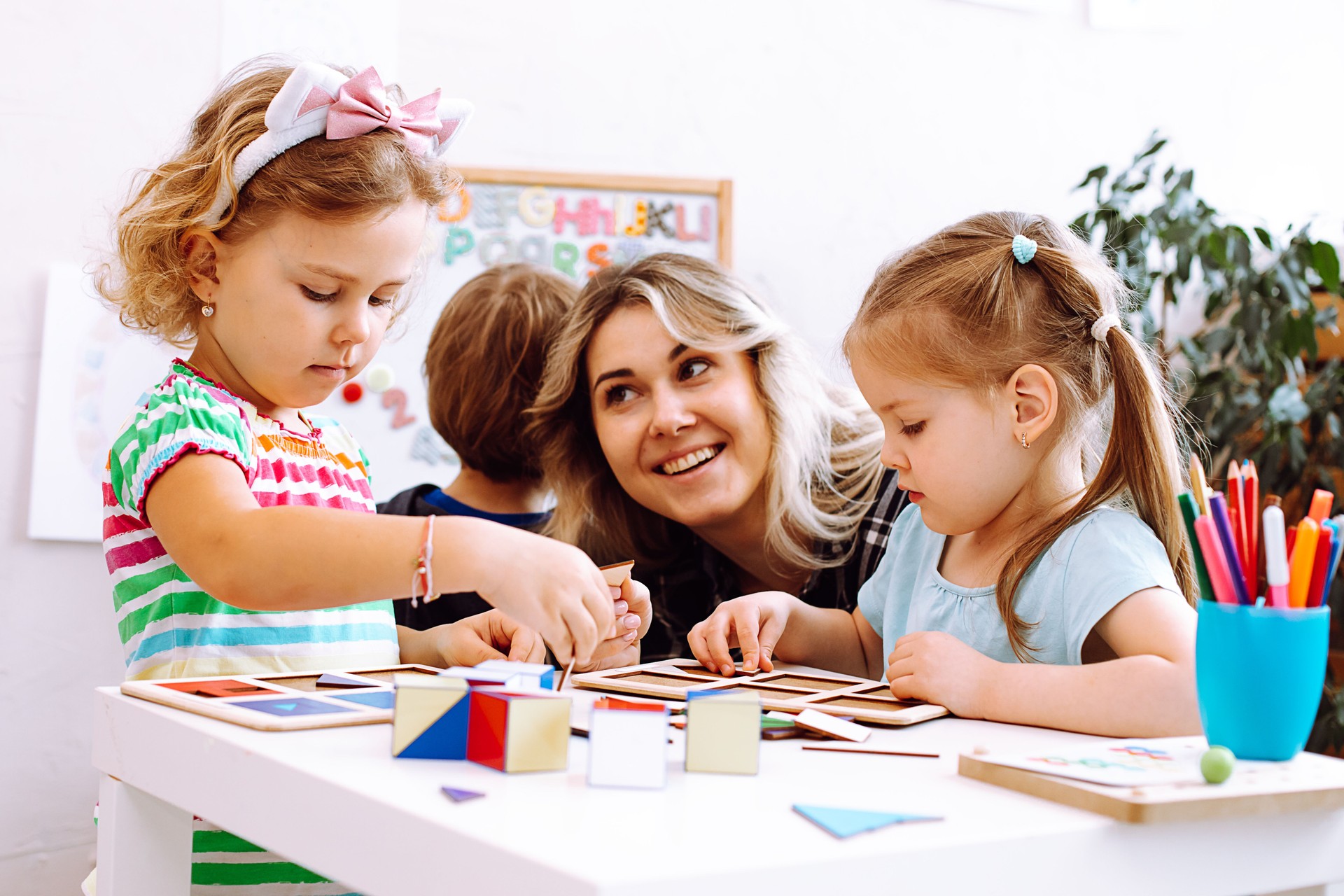 Portrait of young happy woman sitting at table with colored pencils board games. Teacher looking at serious little girl.